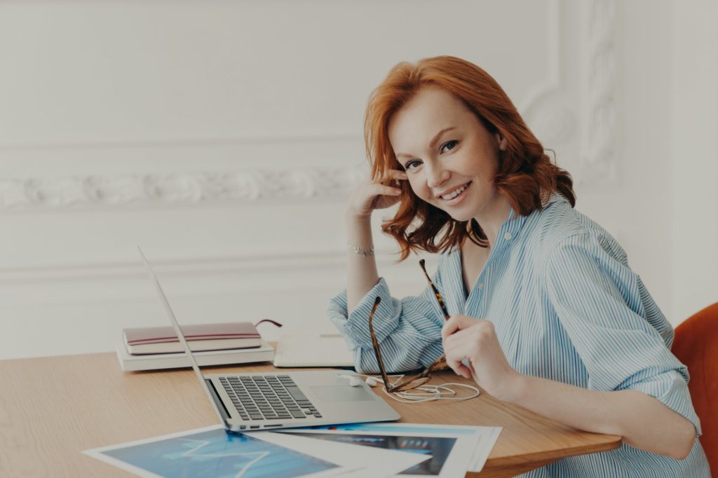 Satisfied businesswoman with foxy hair, checks accounting documentation in online database